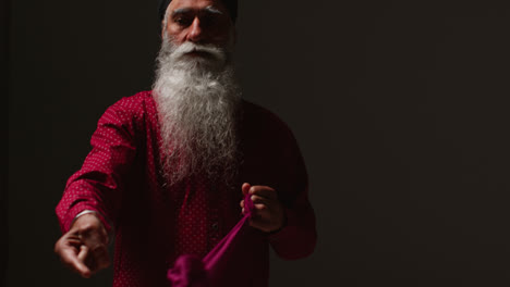 Fotografía-De-Estudio-Con-Iluminación-Tenue-De-Un-Hombre-Sikh-Mayor-Doblando-Tela-Para-Un-Turbante-Contra-Un-Fondo-Oscuro-Liso